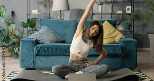 Pretty young Caucasian woman sitting on floor in front of the laptop computer at home and training with online trainer. Attractive slim female working out in iving room with internet video lesson. photo