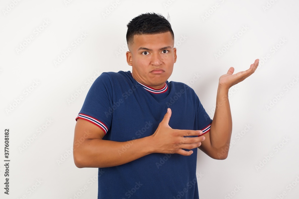 Young handsome hispanic man wearing casual t-shirt standing over white pointing aside with both hands showing something strange and saying: I don't know what is this. Advertisement concept. Young handsome hispanic man wearing casual t-shirt standing over white pointing aside with both hands showing something strange and saying: I don't know what is this. Advertisement concept.