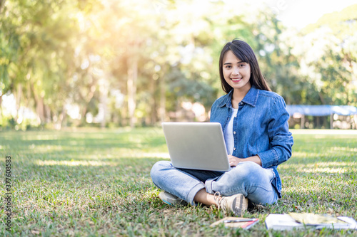 asian student studying outside on filed, on break sitting down on grass filed using laptop computer with books, pencil and note book, studying hard under the trees nature in the evening sunlight