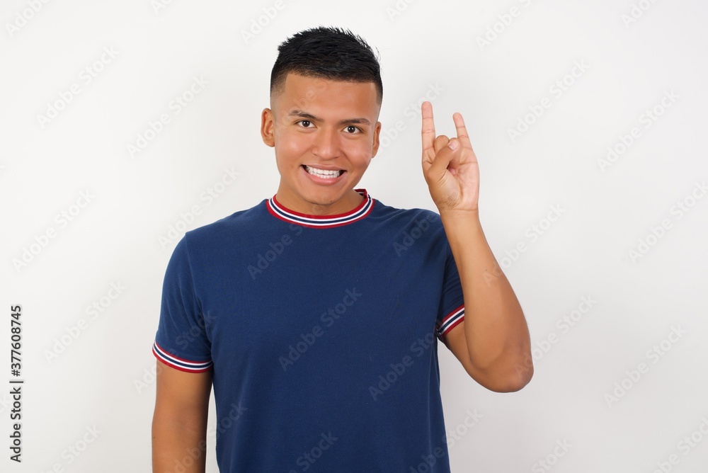 Young handsome hispanic man wearing casual t-shirt standing over white isolated background doing a rock gesture and smiling to the camera. Ready to go to his favorite band concert. Young handsome hispanic man wearing casual t-shirt standing over white isolated background doing a rock gesture and smiling to the camera. Ready to go to his favorite band concert.