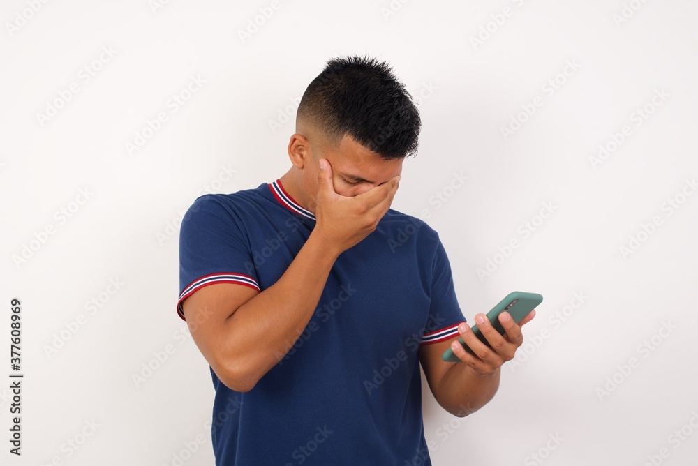 Young handsome hispanic man wearing casual t-shirt standing over white isolated background looking at smart phone feeling sad holding hand on face. Young handsome hispanic man wearing casual t-shirt standing over white isolated background looking at smart phone feeling sad holding hand on face.