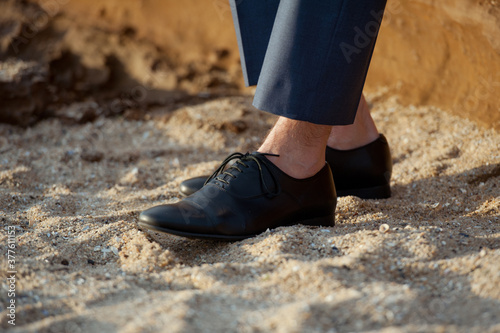 Detail of young handsome man wearing blue suit pants and black leather shoes. He is sitting on the beach on a sunset.