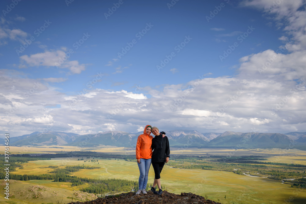 Two young happy girls standing on hill hugging and smile. Women travelers are photographed against the beautiful mountain landscape. Active family recreation and adventure.