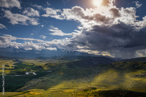 Majestic panorama of mountain plain on the background of snow-covered ridge before thunderstorm. Sun's rays break through huge clouds and beautifully illuminate green steppe and winding river. © exebiche