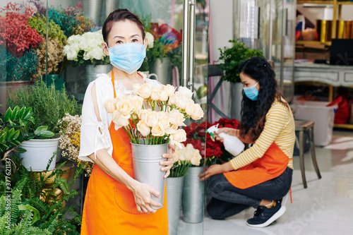 Portrait of senior florist standing in store with metal bucket of white roses  her daughter spraying flowers in background