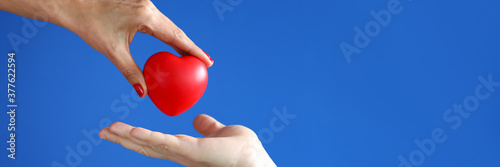 Female hand passes red heart to male hand against blue background closeup. Kindness and charity concept photo