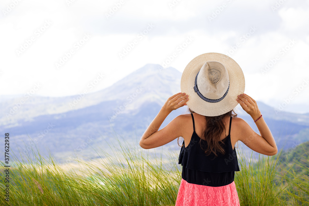 Young traveler woman wearing hat, enjoying mountain landscape. Hills and Batur volcano. Scenic panoramic view. Sky with white clouds. View from back. Copy space. Kintamani, Bali