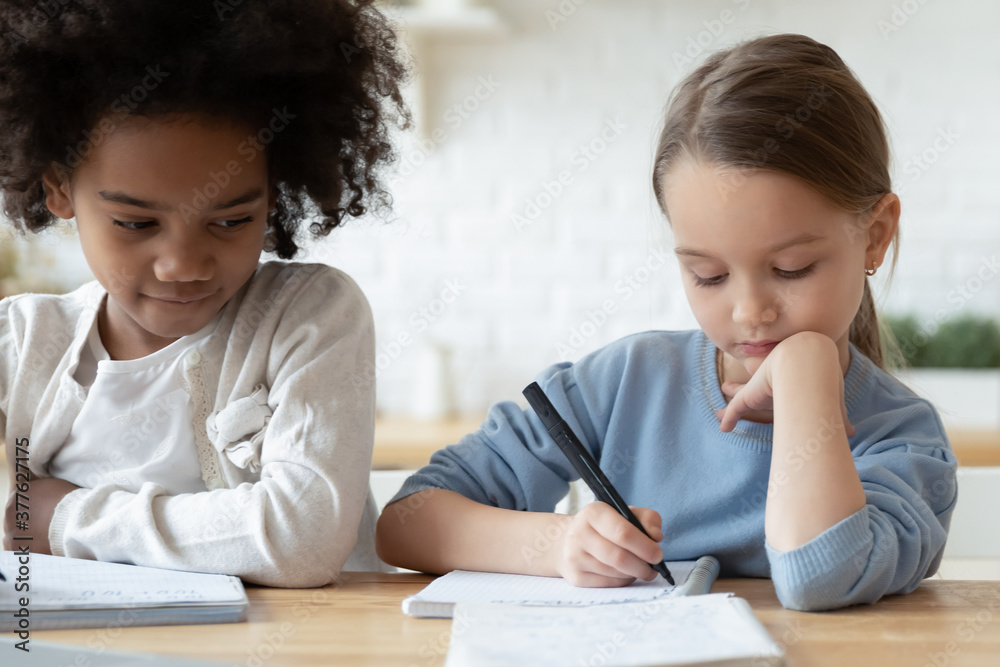 Head shot focused little multiracial sisters sitting at table, preparing homework together indoors. Concentrated adorable 7s small mixed race children girls writing notes, homeschooling concept. Head shot focused little multiracial sisters sitting at table, preparing homework together indoors. Concentrated adorable 7s small mixed race children girls writing notes, homeschooling concept.