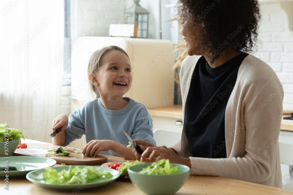 Happy adorable small caucasian kid girl enjoying cooking process with smiling african american babysitter foster mother in modern kitchen, positive multiracial family preparing healthy food together.
