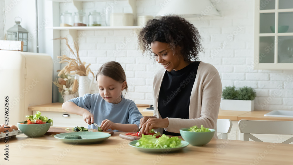 Head shot smiling beautiful african american mother or nanny cooking healthy vegetarian dinner with curious small adorable preschool caucasian kid girl, standing at wooden countertop in modern kitchen