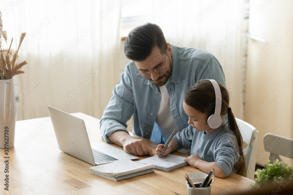 Focused little adorable european girl sitting at desk with wireless headphones, involved in e-learning with caring young father at home. Daddy helping small 7s old kid daughter preparing homework. Focused little adorable european girl sitting at desk with wireless headphones, involved in e-learning with caring young father at home. Daddy helping small 7s old kid daughter preparing homework.