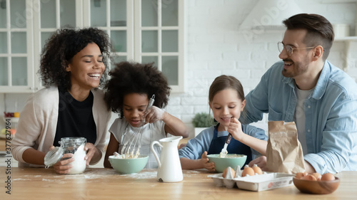 Emotional happy mixed race parents involved in preparing dough for morning breakfast pancakes with little cute biracial daughters in modern kitchen, smiling bonding loving multiethnic family pastime.
