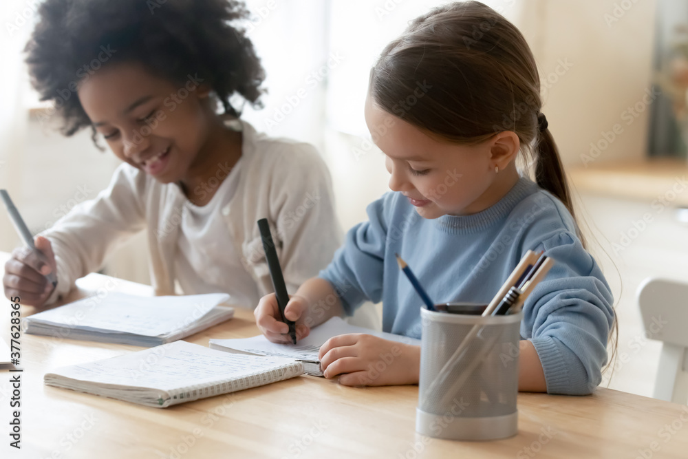 Happy little european kid girl sitting at table with african american foster sister, doing school exercises together at home. Smiling adorable small multiracial children enjoying preparing homework. Happy little european kid girl sitting at table with african american foster sister, doing school exercises together at home. Smiling adorable small multiracial children enjoying preparing homework.