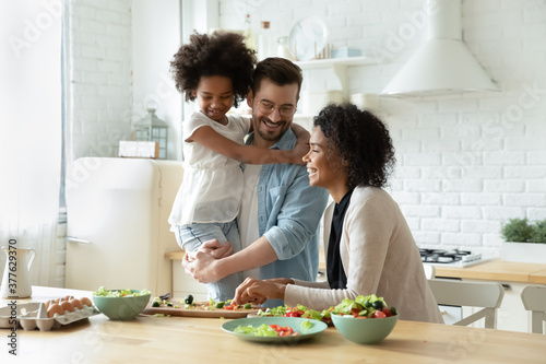 Smiling young european man holding on hands pretty small biracial adopted girl daughter, watching happy african american mother preparing healthy food, chopping fresh vegetables for salad in kitchen.