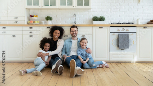 Full length portrait relaxed happy multiracial couple with cute kids sitting on warm wooden floor in modern kitchen. Smiling mixed race family homeowners enjoying peaceful stress free weekend time.