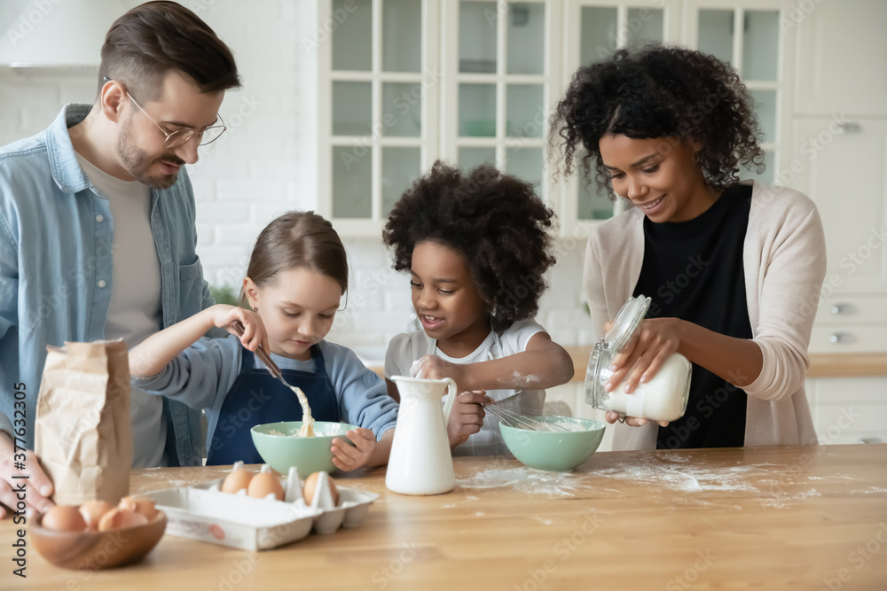 Happy interested multiethnic small children girls in aprons involved in preparing dough for homemade pastry with affectionate loving african american mommy and european father in modern kitchen.... Happy interested multiethnic small children girls in aprons involved in preparing dough for homemade pastry with affectionate loving african american mommy and european father in modern kitchen....