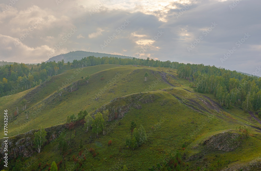 Mountain-hill with rocky areas. Dirt road up. Green forest, birch trees, grass. Dense clouds with sun rays. Mountain landscape concept, hiking, tourism.