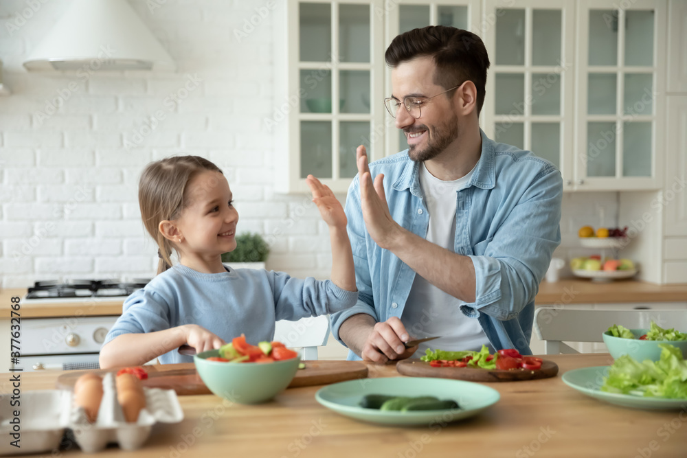 Happy preschool cute small kid girl giving high five to loving caring european father, enjoying cooking together at modern countertop, positive family celebrating preparing healthy food dinner. Happy preschool cute small kid girl giving high five to loving caring european father, enjoying cooking together at modern countertop, positive family celebrating preparing healthy food dinner.