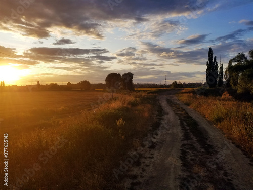 dirt road along a mown field in the rays of sunset, from above there are large clouds in the sky