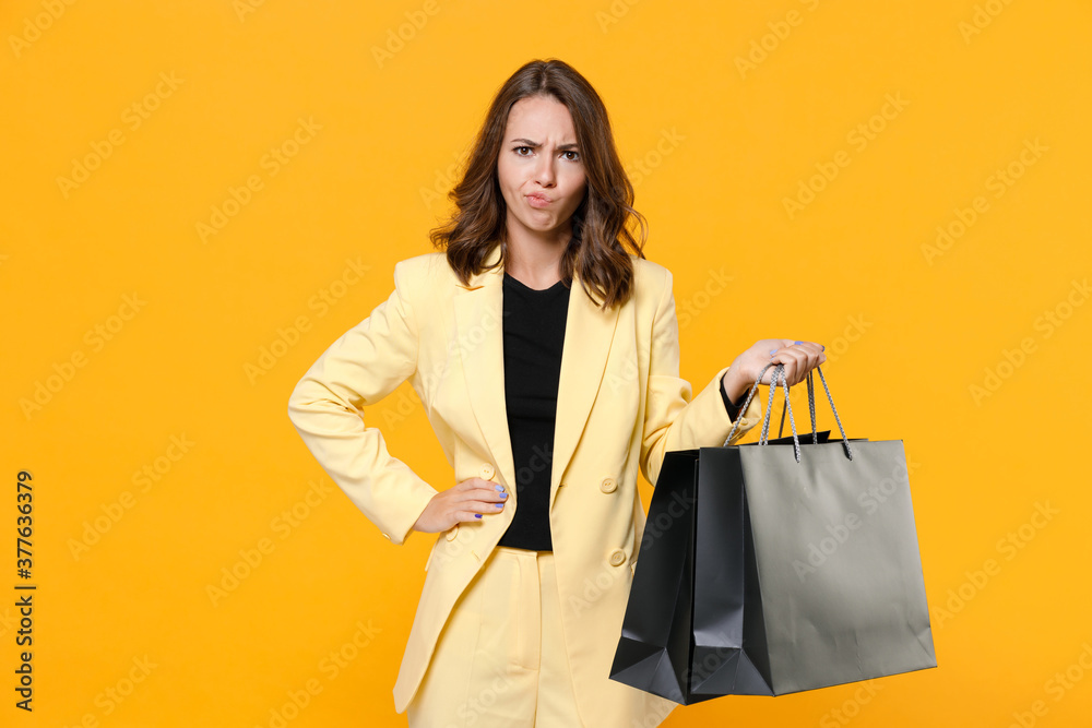 Displeased confused puzzled young woman 20s in basic light suit jacket hold package bags with purchases after shopping looking camera isolated on yellow background studio portrait. Black friday sale. Displeased confused puzzled young woman 20s in basic light suit jacket hold package bags with purchases after shopping looking camera isolated on yellow background studio portrait. Black friday sale.