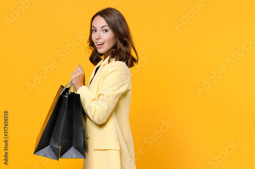 Side view of amazed young brunette woman 20s in basic light suit jacket hold package bags with purchases after shopping looking camera isolated on yellow background studio portrait. Black friday sale.