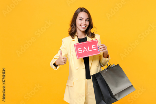Smiling young woman 20s in basic light suit jacket hold package bags with purchases after shopping sign with SALE title showing thumb up isolated on yellow background, studio portrait. Black friday.