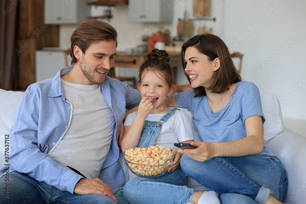 Happy family with child sitting on sofa watching tv and eating popcorn, young parents embracing daughter relaxing on couch together.