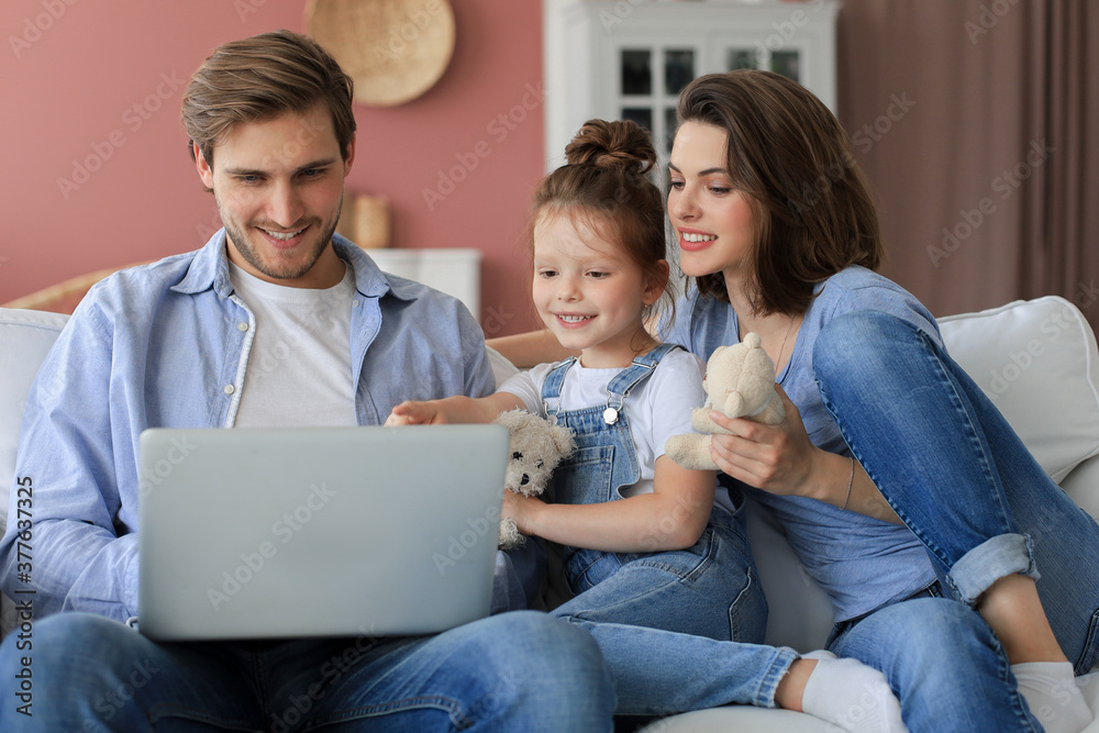 Positive friendly young parents with smiling little daughter sitting on sofa together answering video call on laptop and waving hand in greeting while relaxing at home on weekend Positive friendly young parents with smiling little daughter sitting on sofa together answering video call on laptop and waving hand in greeting while relaxing at home on weekend