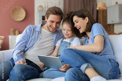 Positive friendly young parents with smiling little daughter sitting on sofa together answering video call on digital tablet while relaxing at home on weekend.