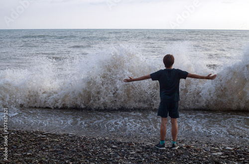 Boy standing on the seashore with outstretched arms against the backdrop of stormy waves.
