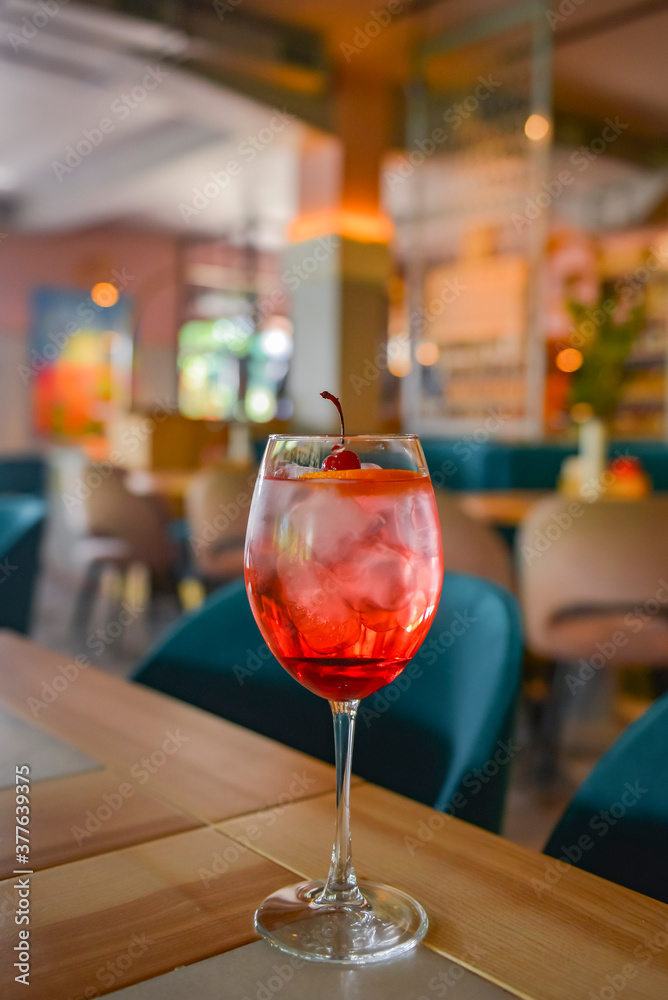 Red cocktail with ice served in a glass on wooden table with blurred restaurant background. Refreshing summer beverage.