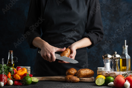 Chef cleans potatoes on the restaurant's kitchen. For dishes with potatoes.. Some fruits and vegetables on the table. Girls hands with knife. Healthy meal. Dark background.