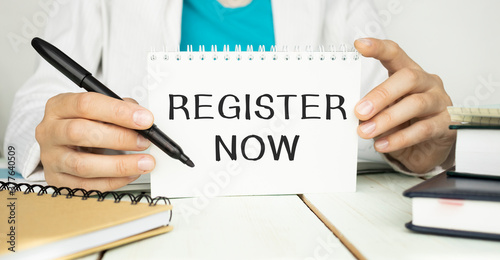 Register now is written in a notebook in the hands of a businessman.