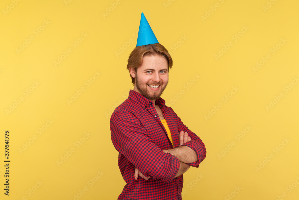 Happy guy with funny party cone on head standing crossed hands, looking at camera with toothy smile, celebrating birthday holiday, anniversary. indoor studio shot isolated on yellow background