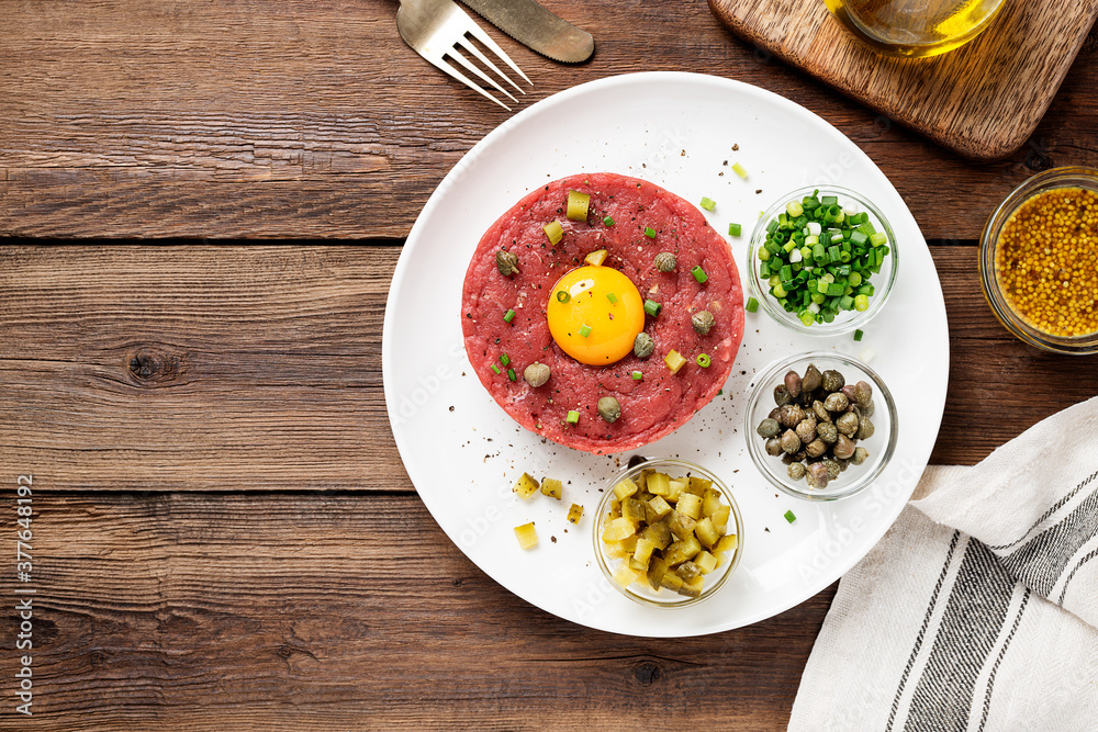 Beef tartare with capers , fresh onion and pickled cucumber on white plate on wooden background. top view