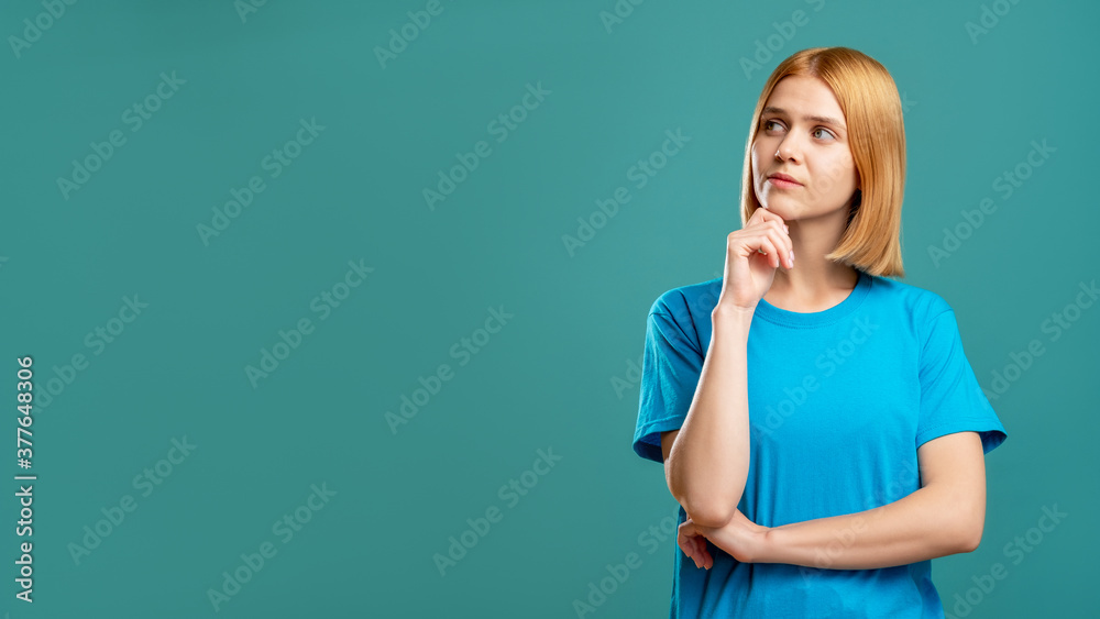 Curious woman portrait. Special offer. Thoughtful doubtful blonde lady in blue t-shirt looking at copy space for brand text isolated on teal commercial background. Hard choice. Curious woman portrait. Special offer. Thoughtful doubtful blonde lady in blue t-shirt looking at copy space for brand text isolated on teal commercial background. Hard choice.