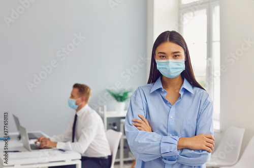 Serious young woman wearing face mask standing in her workplace looking at camera