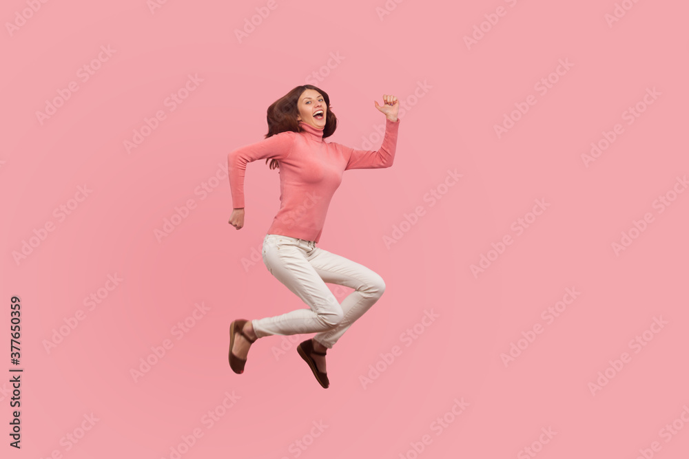 Extremely happy satisfied woman with brown hair jumping high or flying in air, dreams comes true. Indoor studio shot isolated on pink background