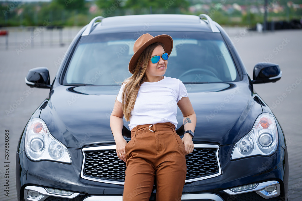 A beautiful girl of European appearance with glasses and a brown hat is standing near a black car. Young woman with car in parking A beautiful girl of European appearance with glasses and a brown hat is standing near a black car. Young woman with car in parking