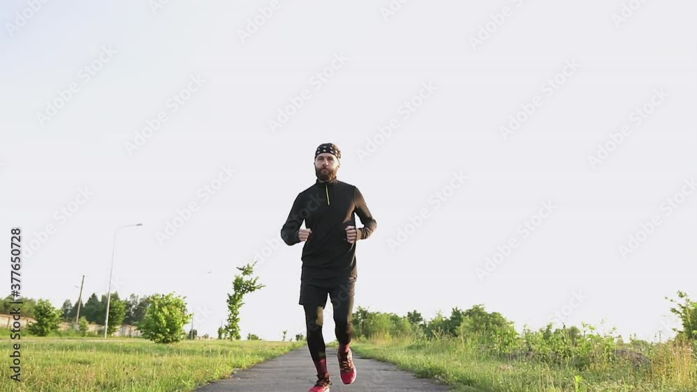 Close up of handsome confident strong bearded young athlete in black clothes which running on the pathway near green field Close up of handsome confident strong bearded young athlete in black clothes which running on the pathway near green field