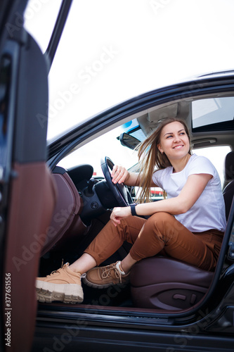 Young beautiful woman with long hair sits in a black car at a parking lot. Pretty girl in casual clothes. Car trip