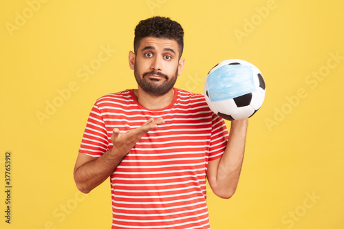 Disappointed bearded man in red t-shirt showing soccer ball in safety mask, sport competition during coronavirus epidemic. Indoor studio shot isolated on yellow background