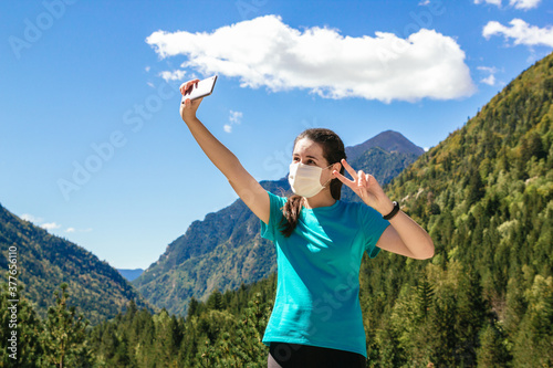 Stock photo of a 30's woman with face mask taking a selfie with her phone while enjoying the mountains on a sunny day