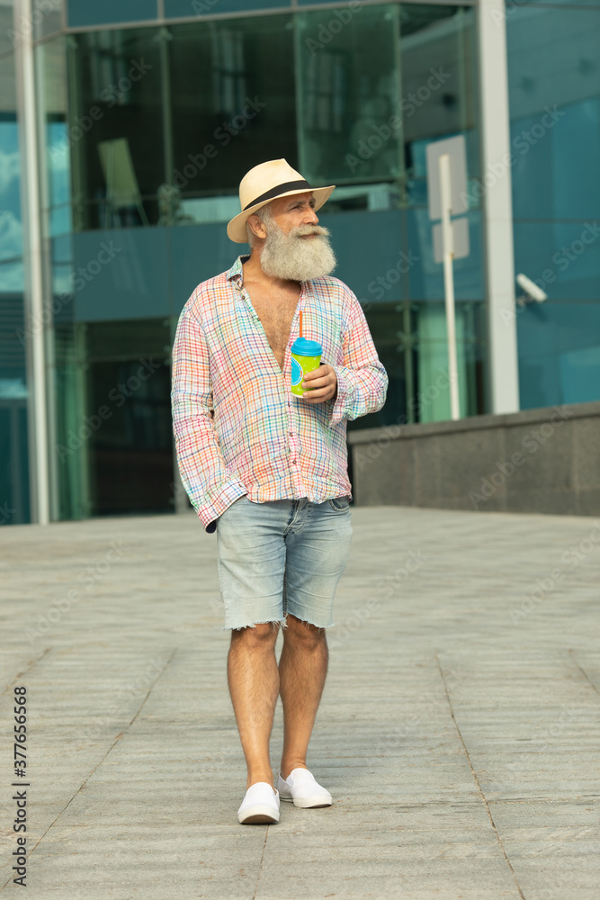 Portrait of positive senior bearded hipster man having coffee break outdoors on urban settings.