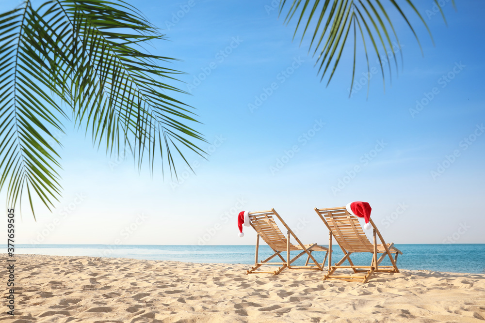 Sun loungers with Santa's hats on beach, space for text. Christmas vacation - obrazy, fototapety, plakaty 