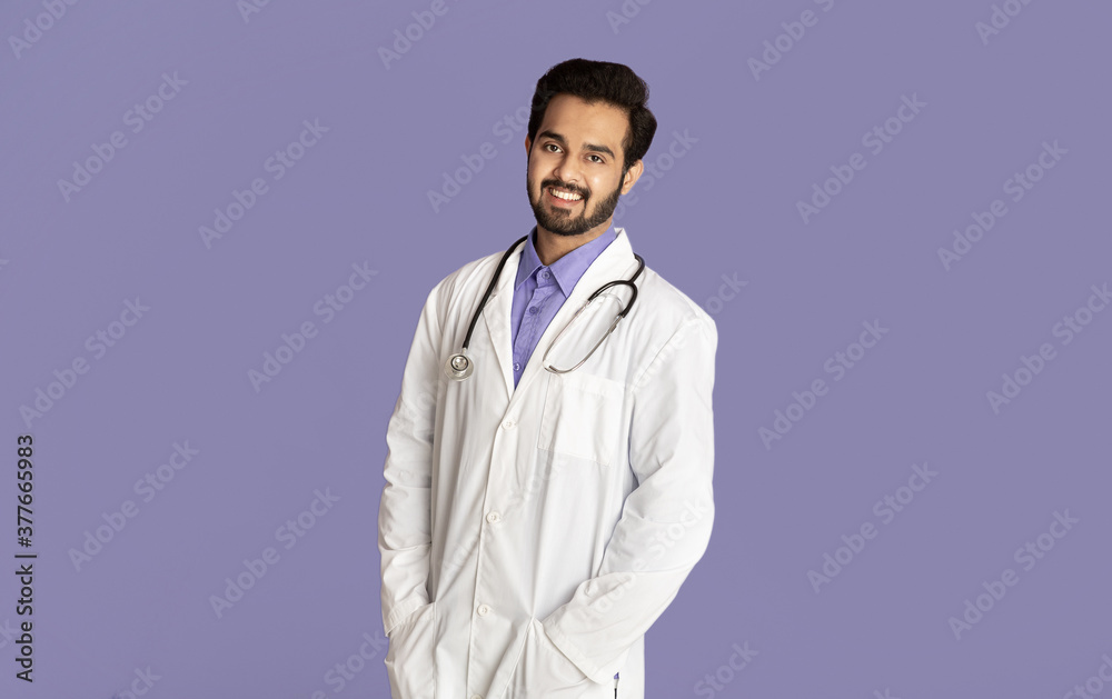 Portrait of confident Indian medical doctor in lab coat looking at camera on violet background Portrait of confident Indian medical doctor in lab coat looking at camera on violet background