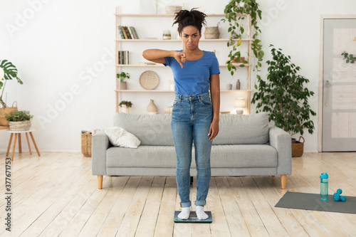 Frustrated Black Girl Gesturing Thumbs-Down Standing On Scales Indoors