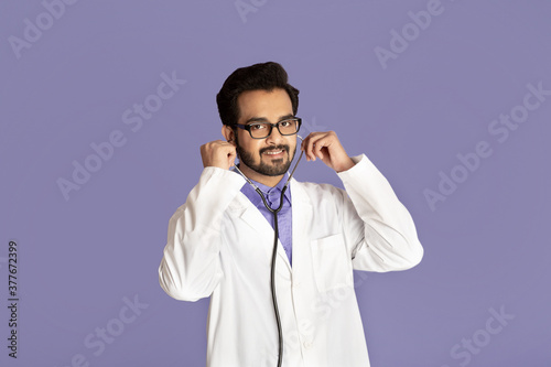 Portrait of cheerful Indian doctor in lab coat with stethoscope on violet background