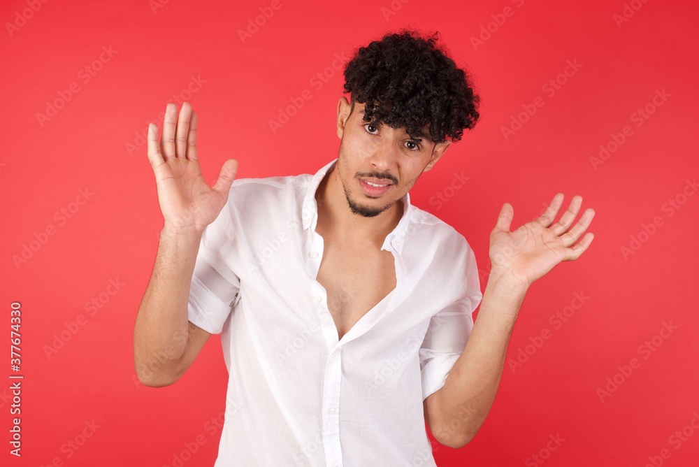 Clueless Young arab man with afro hair wearing shirt standing over isolated red background shrugs shoulders with hesitation, faces doubtful situation, spreads palms, Hard decision Clueless Young arab man with afro hair wearing shirt standing over isolated red background shrugs shoulders with hesitation, faces doubtful situation, spreads palms, Hard decision