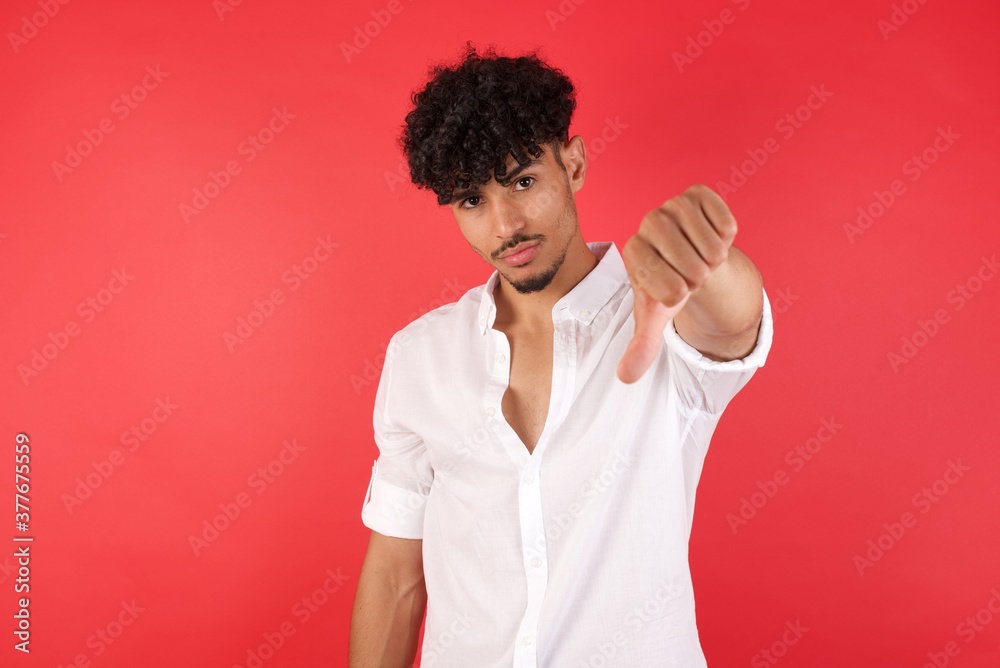 Young arab man with afro hair wearing shirt standing over isolated red background feeling angry, annoyed, disappointed or displeased, showing thumbs down with a serious look Young arab man with afro hair wearing shirt standing over isolated red background feeling angry, annoyed, disappointed or displeased, showing thumbs down with a serious look